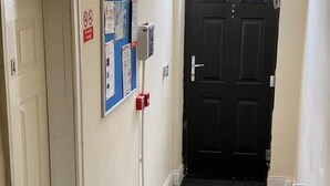 A narrow hallway with a carpeted floor, a door on the left, and a black door with a fire exit sign on the right. - House 2 Bedrooms Sleeps 4 With Parking (Bolton)