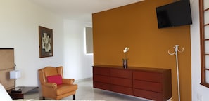 Ein Hotelzimmer mit einem braunen Sofa, einem roten Schminktisch, einem an der Wand montierten Fernseher und einem gerahmten Kunstwerk.