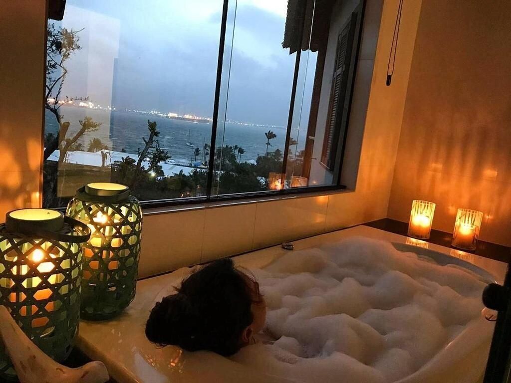 Uma banheira cheia de bolhas, uma janela com vista para o mar e duas velas acesas ao lado.