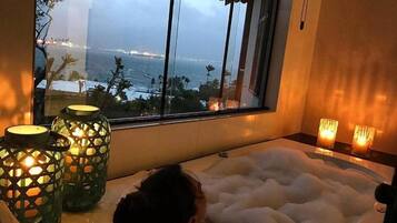 Uma banheira cheia de bolhas, uma janela com vista para o mar e duas velas acesas ao lado.