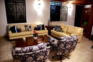 Una sala de estar con dos sofás, una mesa de centro con un jarrón de flores, sillones estampados y apliques decorativos en la pared.