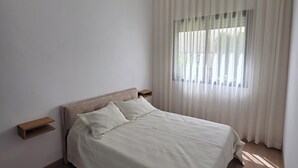 Un dormitorio moderno con cama, cabecera de madera, mesitas de noche y un ventanal con cortinas traslúcidas.
