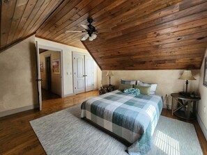 Ein Schlafzimmer mit schrägverdeckter Decke, einem Bett mit kariertem Überwurf, zwei Nachttischen mit Lampen, einer Tür zu einem anderen Raum und einem kleinen runden Tisch mit Lampe und Pflanzen.