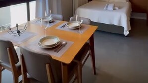 A hotel room with a bed, a dining table with chairs, and a view of a building outside. - Fully Equipped Studio Apartment (Joinville)