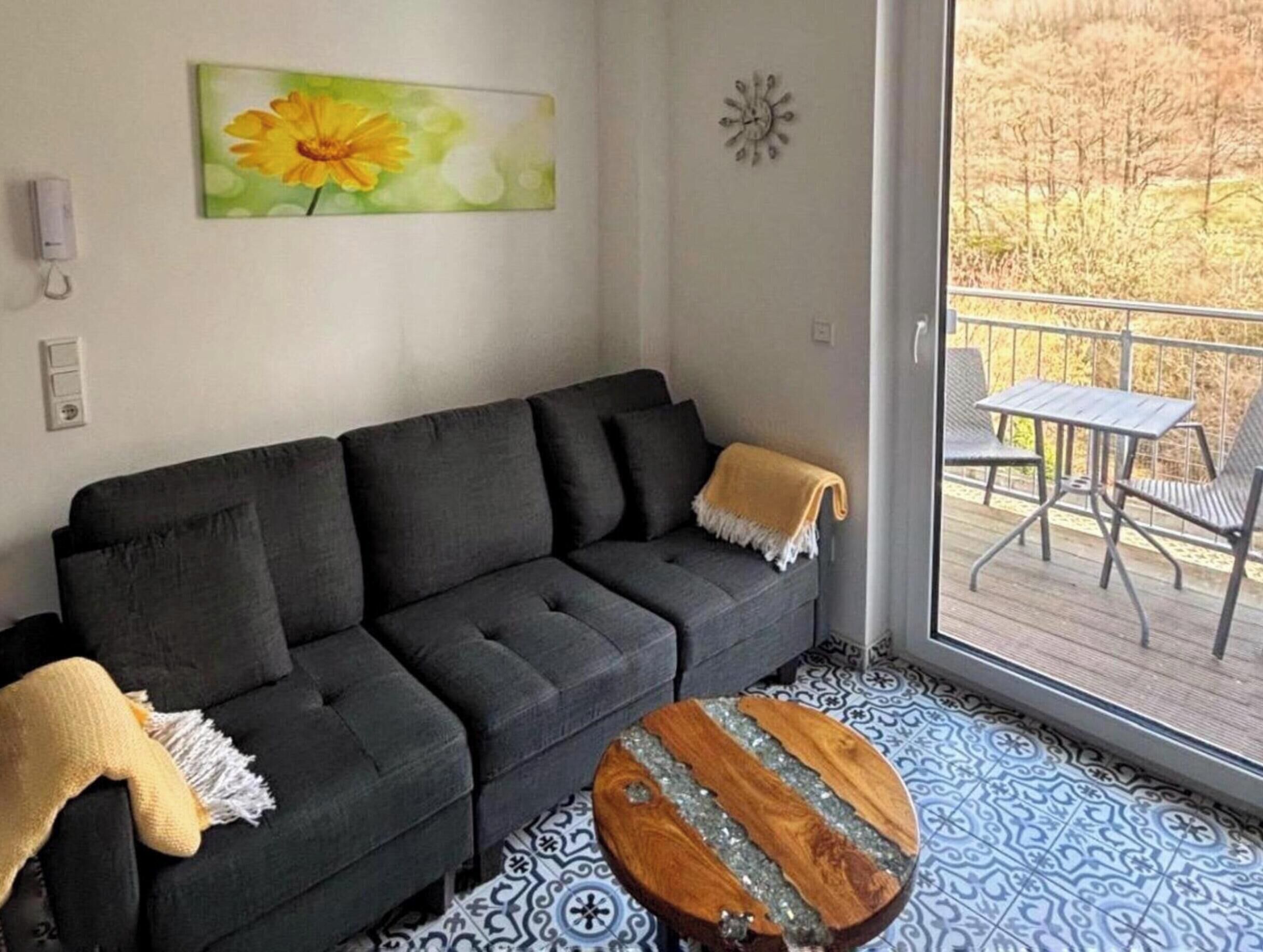 A modern living room with a grey sofa, a wooden coffee table, and a large window offering a view of the outdoors.