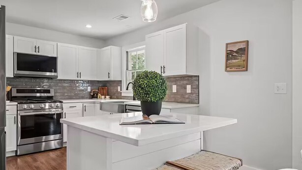 Une cuisine moderne avec des meubles blancs, des appareils en acier inoxydable, un îlot central agrémenté d’une plante en pot et un luminaire suspendu.