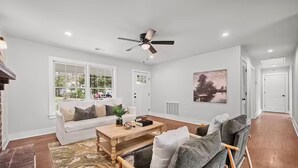 Un salon avec un canapé blanc, une table basse en bois, un fauteuil gris, un ventilateur de plafond et une grande fenêtre donnant sur l’extérieur.