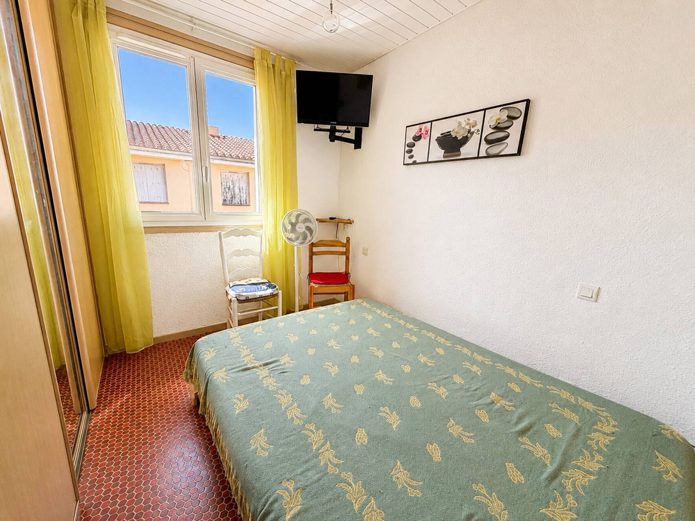 Una habitación con cama, una silla, ventilador, televisor y ventana con cortinas.