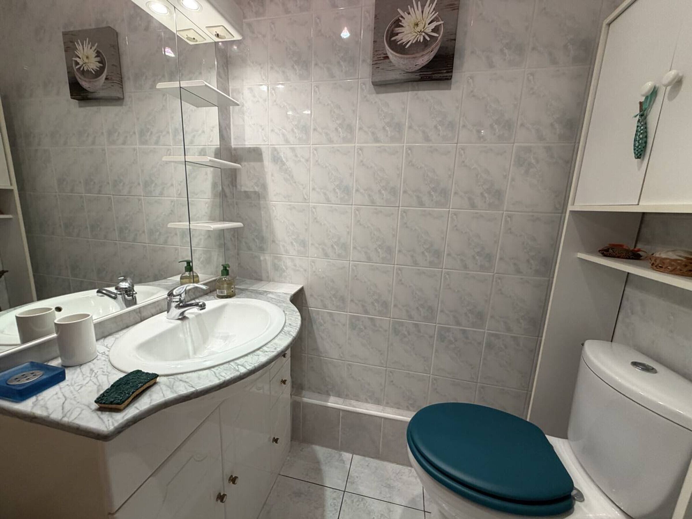 Ein Badezimmer mit Waschbecken, Spiegel und Toilette. Zwei Ablagen mit Pflegeprodukten und eine blaue Matte liegen auf dem Boden.