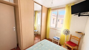 Quarto com cama, cadeira, ventoinha, televisão e janela com cortinas.