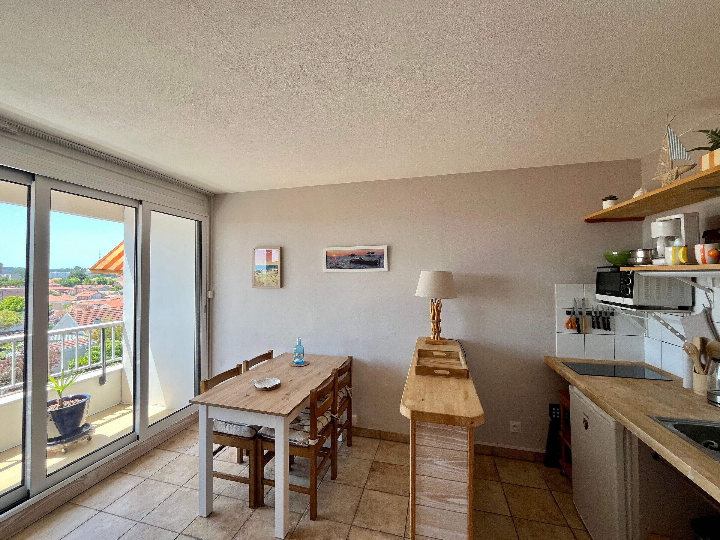 Ein Balkon mit Blick auf Gebäude, ein Esstisch mit Stühlen, ein kleiner Tisch mit Lampe sowie eine Küchenzeile mit Mikrowelle und Geschirr.