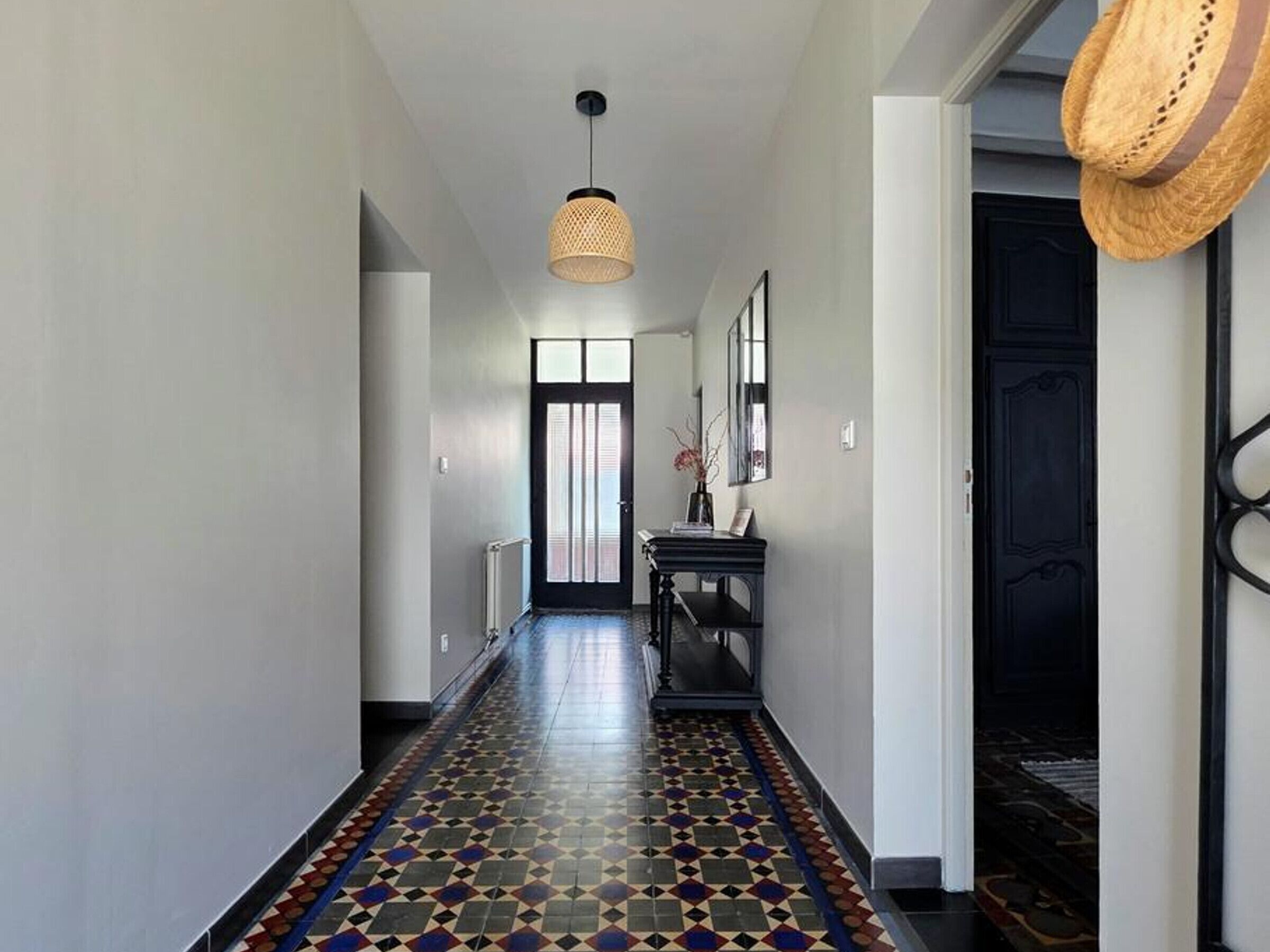 A hallway with patterned flooring, a black door, a hanging light fixture, and a wall-mounted mirror.