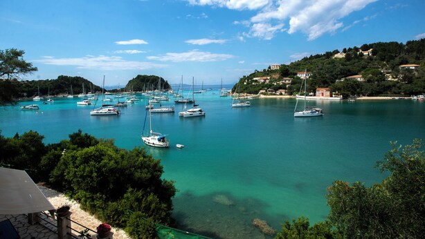 A coastal marina with multiple boats docked, surrounded by lush greenery and hills in the background. - Villa Praxithea (Lakka)