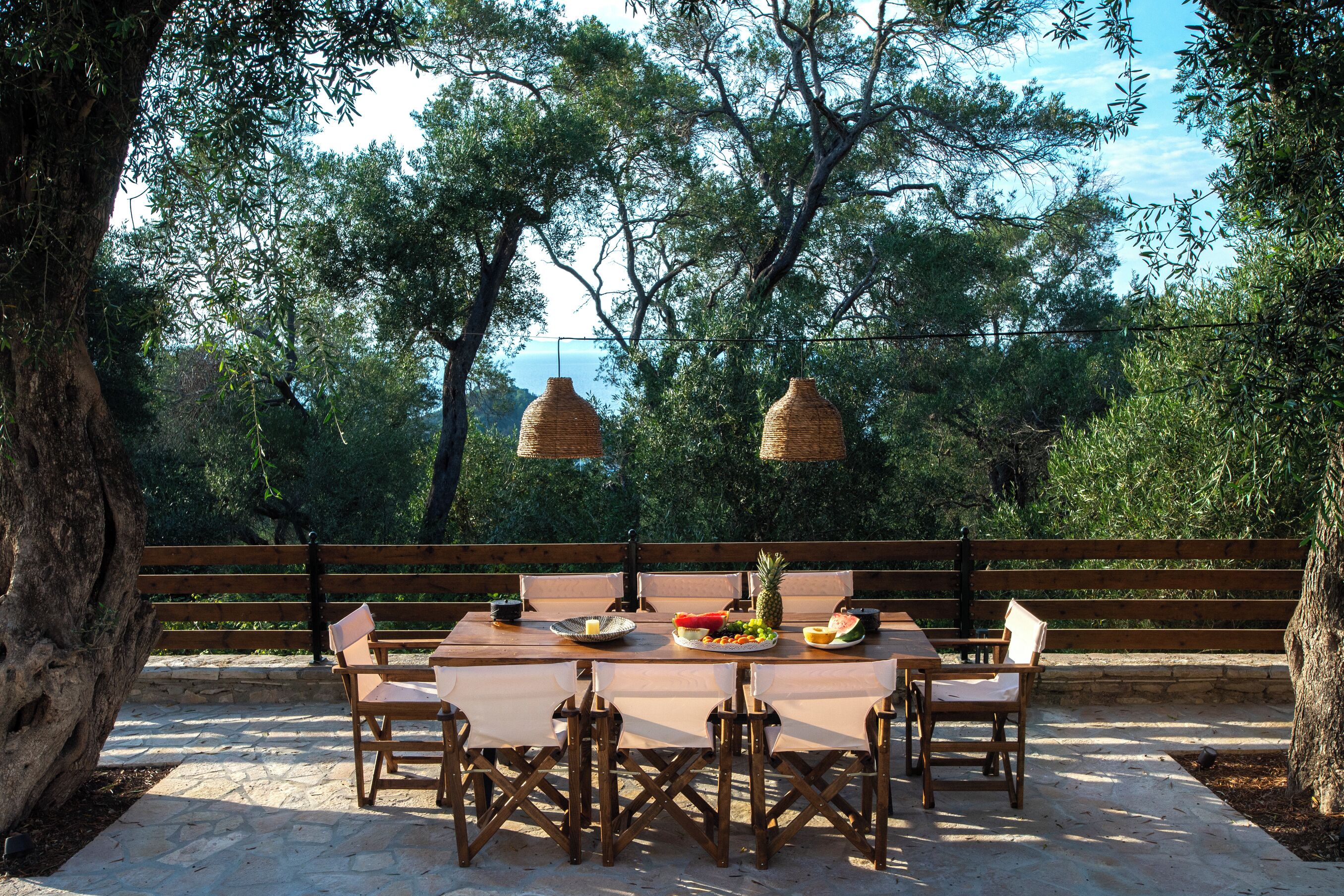 An outdoor dining area with a stone floor, wooden table and chairs, and hanging wicker lamps.