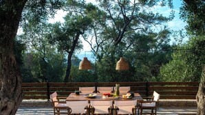 An outdoor dining area with a stone floor, wooden table and chairs, and hanging wicker lamps. - Villa Praxithea (Lakka)