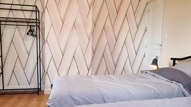 Une chambre avec un mur orné d’un motif en chevrons, un portant métallique avec des vêtements, une lampe de chevet et un plancher en bois.