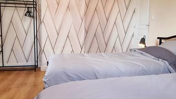 Une chambre avec un mur orné d’un motif en chevrons, un portant métallique avec des vêtements, une lampe de chevet et un plancher en bois.