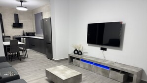 Ein modernes Wohnzimmer mit Flachbildfernseher, grauen Sofas, einem Couchtisch und einer Küchenzeile inklusive Kühlschrank und Mikrowelle.