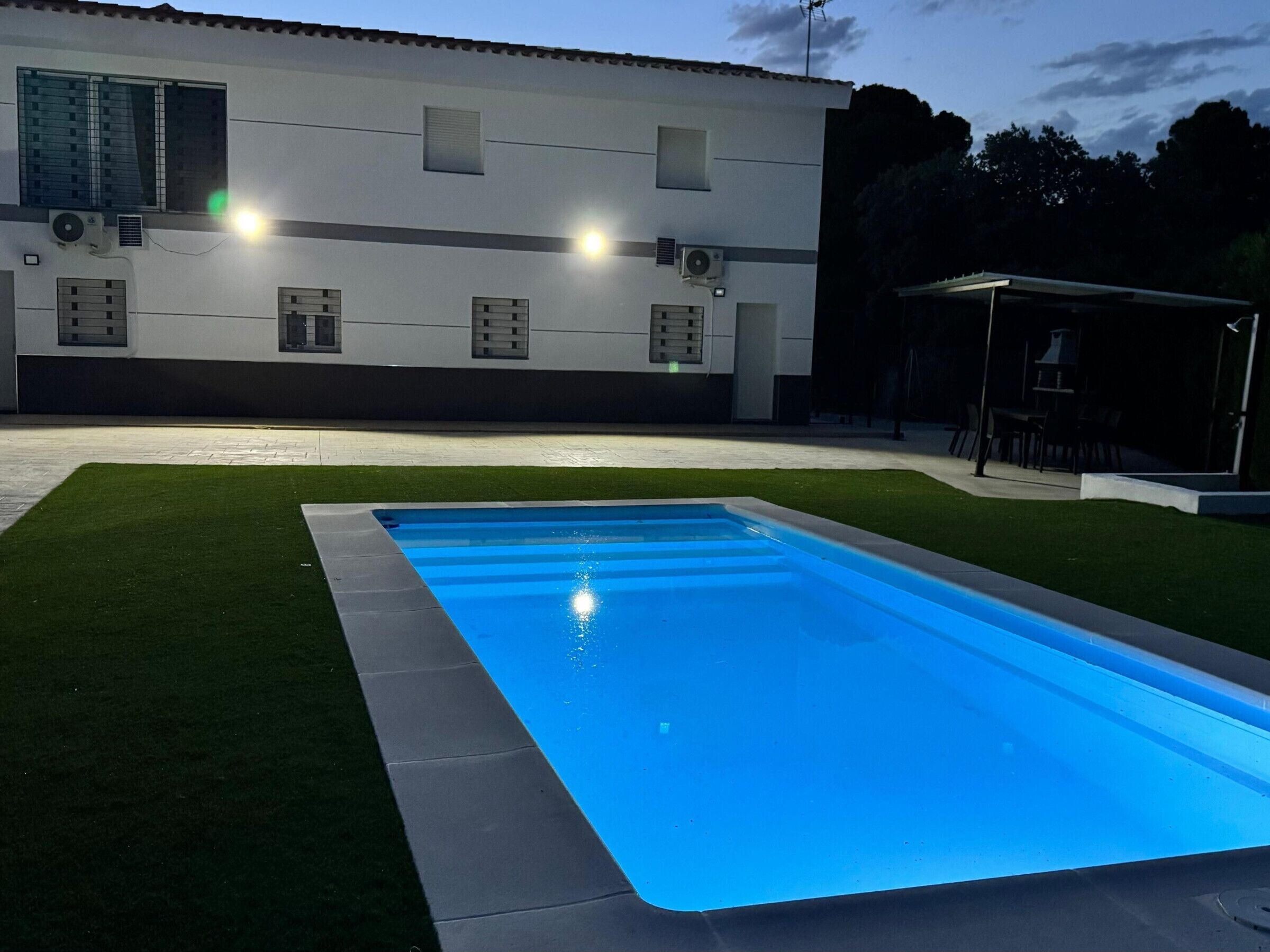 A swimming pool with a tiled edge, surrounded by a grassy area, and a building with multiple windows and air conditioning units in the background.