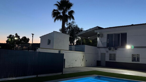 Ein Schwimmbecken mit klarer, blauer Wasseroberfläche, ein weißes Gebäude mit Balkon und eine Palme im Hintergrund.