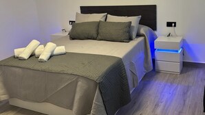 Ein ordentlich gemachtes Bett mit grau-weißer Daunendecke, zwei Kissen und aufgerollten weißen Handtüchern an einem grauen Kopfteil. Ein Nachttisch mit Lampe und Steckdose ist vorhanden.