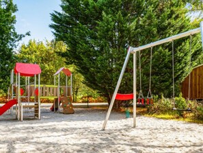 Ein Spielplatz mit Schaukeln und Rutschen, umgeben von Bäumen und einer Holzkonstruktion.