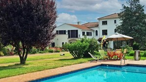 Una piscina con borde de ladrillo, césped y un edificio con tejado de baldosas al fondo.