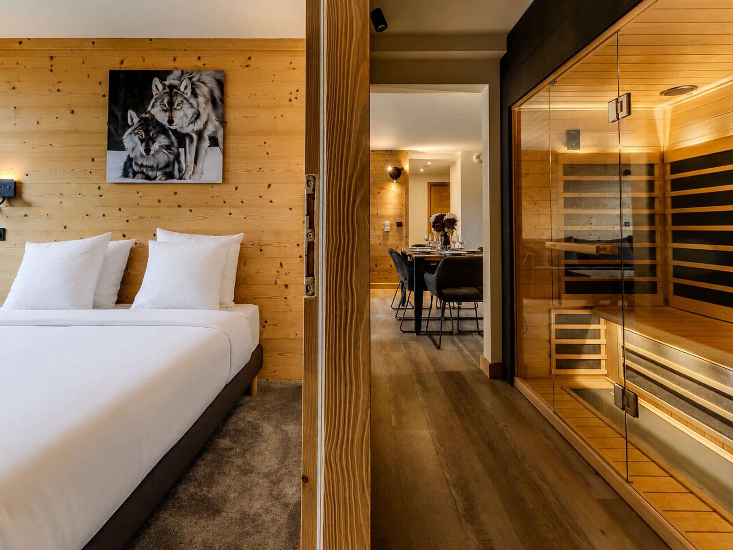 Une chambre d’hôtel avec une tête de lit en bois, un lit avec du linge de maison blanc, un coin repas avec une table et des chaises, et un espace sauna avec des bancs en bois.