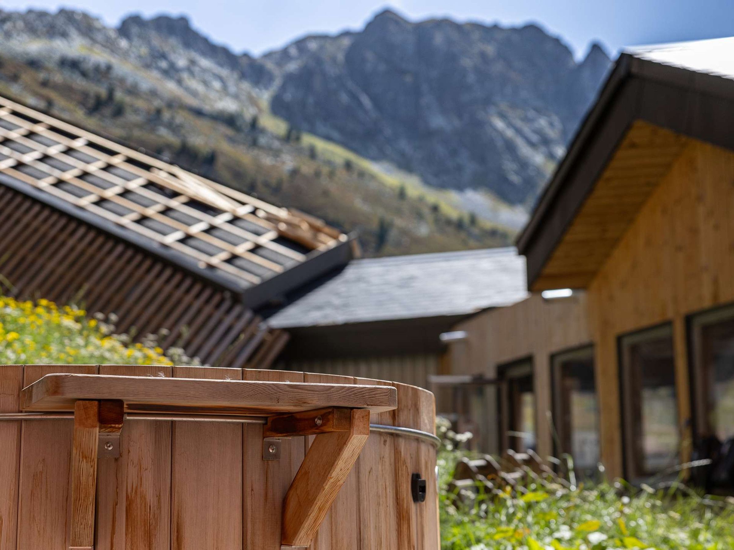 Jacuzzi de madera con estructura metálica, con vistas a montañas y un edificio de madera con ventanas grandes.