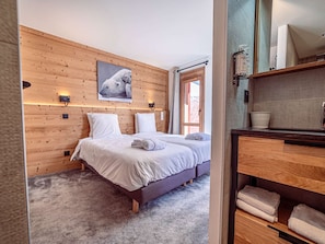 A bedroom with a wooden wall, a bed with white bedding, a window, and a bathroom with a wooden cabinet and a mirror. - Luxury apartment in center - 6 guests (Belle Plagne)