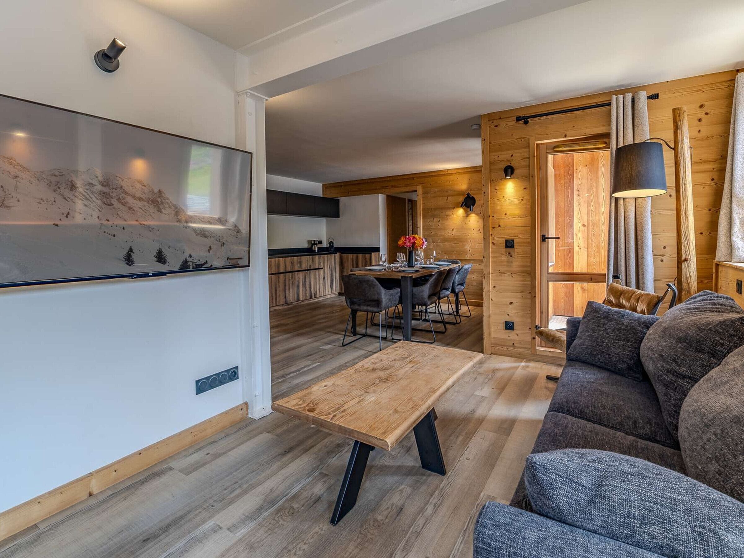 A modern interior with a wooden dining table, a sofa, and a kitchen area visible in the background.