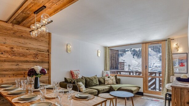 Un tavolo da pranzo in legno apparecchiato con piatti e bicchieri, un divano verde e vista su un paesaggio innevato attraverso un'ampia finestra.