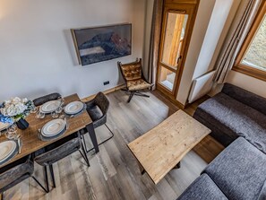 Ein Essbereich mit Holztisch, gedeckt für vier Personen, einer Couch, einem an der Wand befestigten Fernseher und einer Tür, die nach draußen führt.