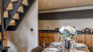 Un espace de salle à manger moderne avec une table en bois dressée pour quatre personnes, un escalier et une cuisine en arrière-plan.