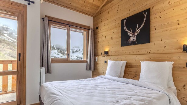Une pièce aux murs lambrissés, comprenant un lit, une fenêtre avec des rideaux, un luminaire fixé au mur et un tableau représentant une tête de cerf.