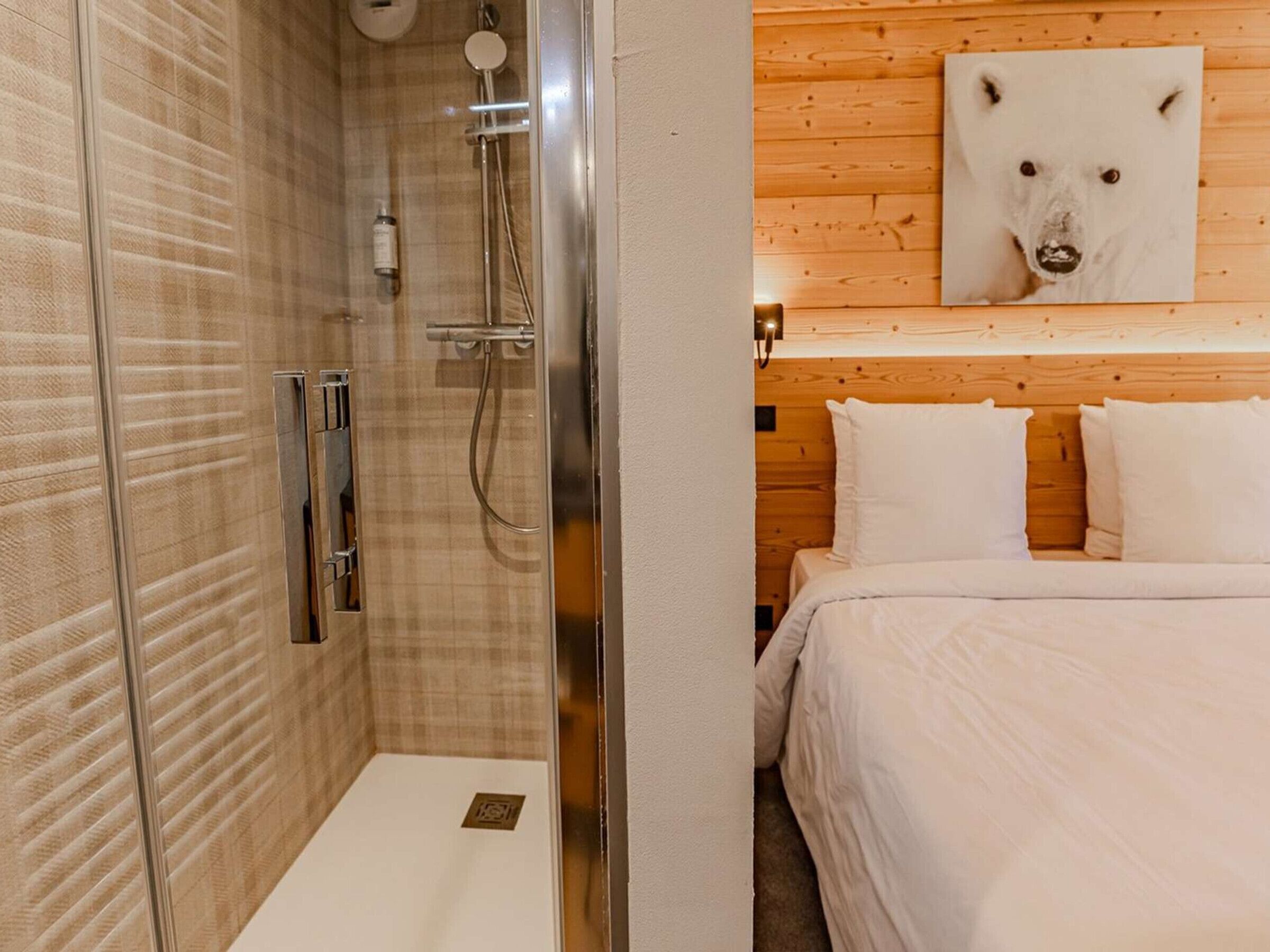 Un baño con cabina de ducha de vidrio, pared de madera y cama con ropa blanca.