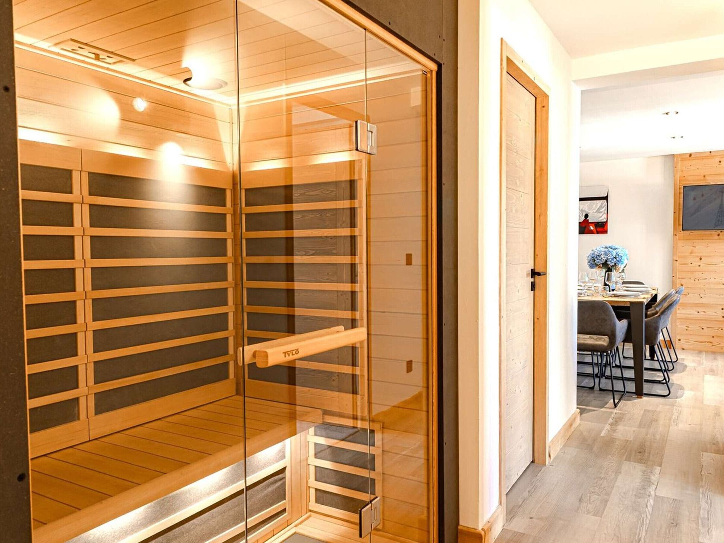 Sauna, jossa on puupanelointi ja lasiovet, ruokailutila pöytineen ja tuoleineen sekä puulattia.