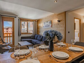 A dining area with a wooden table set for a meal, a sofa, and a window with a view of buildings. - Spacious apartment, 8-10 guests, in Belle Plagne (Belle Plagne)