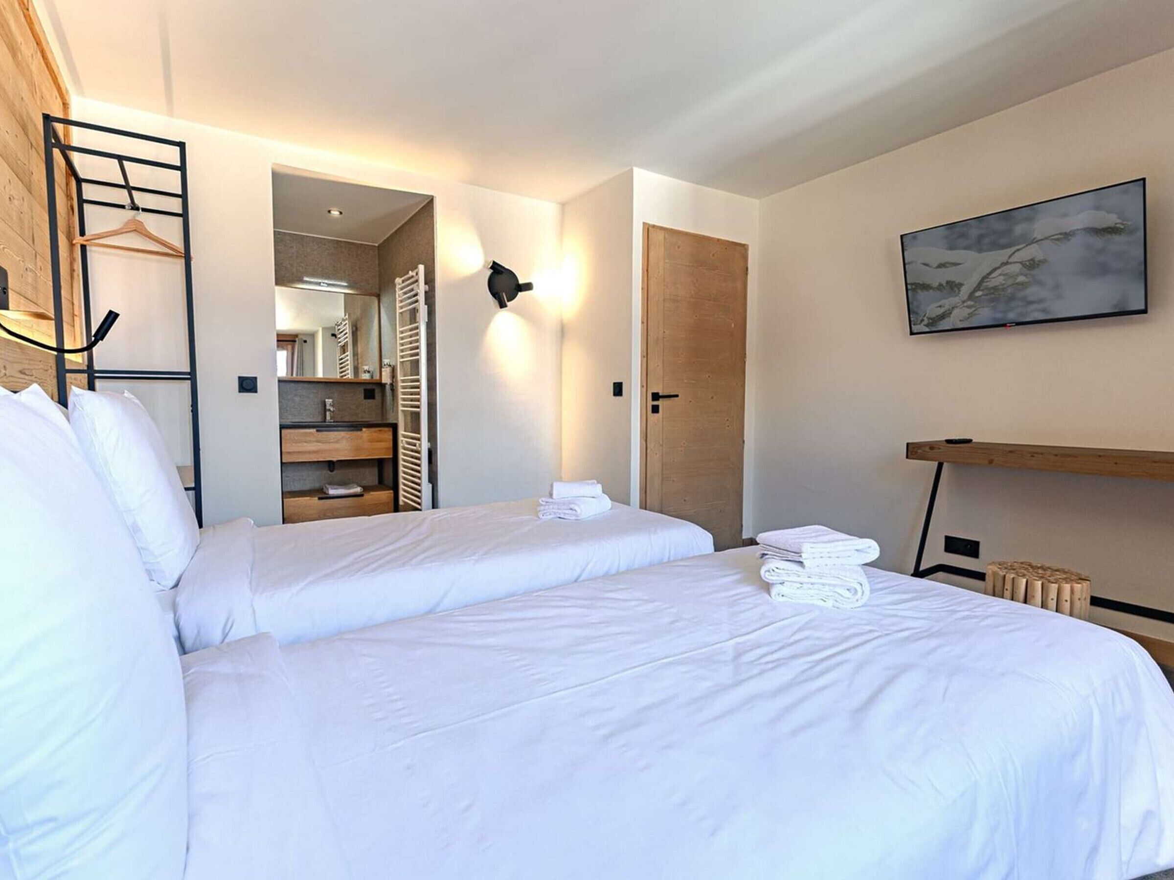 A hotel room with two beds, a wooden wardrobe, a mirror, a towel rack, and a painting on the wall.