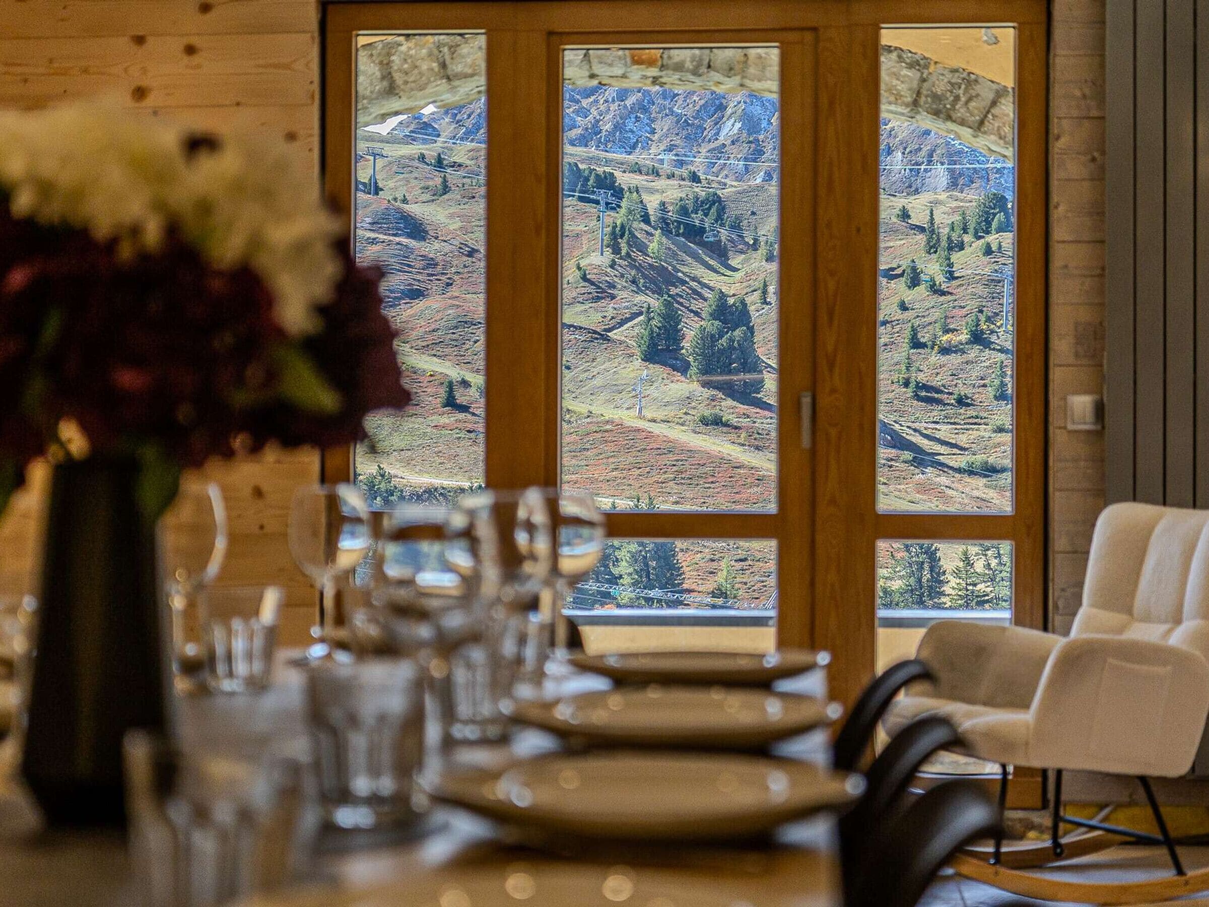 A dining area with a table set for four, a vase with flowers, and a view of a mountainous landscape through a wooden door.