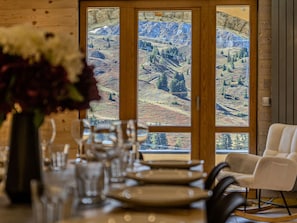 Un comedor con una mesa lista para cuatro, un florero con flores y vista a un paisaje montañoso a través de una puerta de madera.