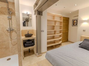 Une salle de bain moderne avec une douche, un lavabo et un miroir. On y trouve un lit avec une couverture et des oreillers gris, ainsi qu’une étagère en bois.