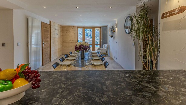 Een eetkamer met een lange tafel gedekt voor een maaltijd, een fruitschaal ernaast en een uitzicht op de omgeving door grote ramen.
