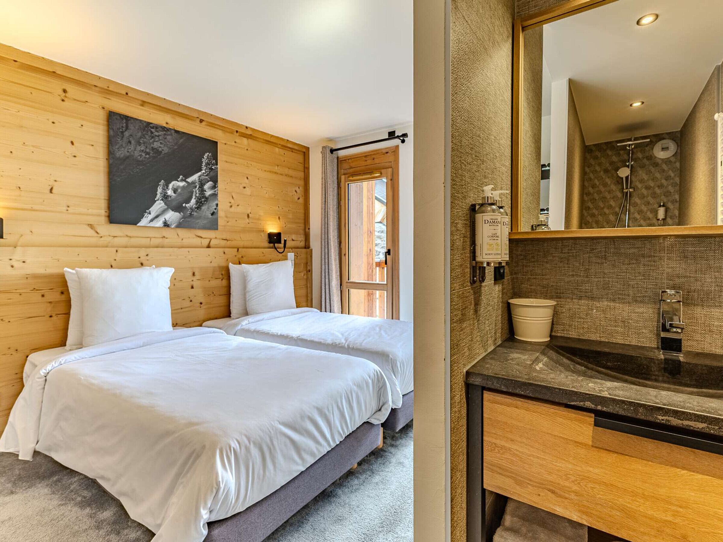 Een hotelkamer met twee bedden, een houten wand, een schilderij en een badkamer met een wastafel en spiegel.
