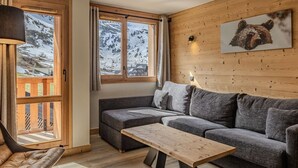 A cozy room with a wooden wall, a gray sofa, a wooden table, and a window with a view of snow-covered mountains. - Cozy apartment - 6 guests, near slopes (Belle Plagne)