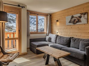 A cozy room with a wooden wall, a gray sofa, a wooden table, and a window with a view of snow-covered mountains. - Cozy apartment - 6 guests, near slopes (Belle Plagne)