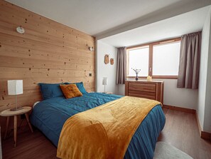 Ein Schlafzimmer mit einem hölzernen Kopfteil, einem Bett mit blau-gelber Bettwäsche, einem Nachttisch mit einer Lampe, einem Schminktisch und einem Fenster mit Vorhängen.