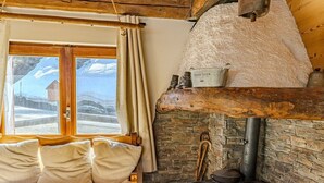 Een rustieke kamer met een houten bank, een stenen muur, een raam met uitzicht op besneeuwde bergen en een houtkachel.