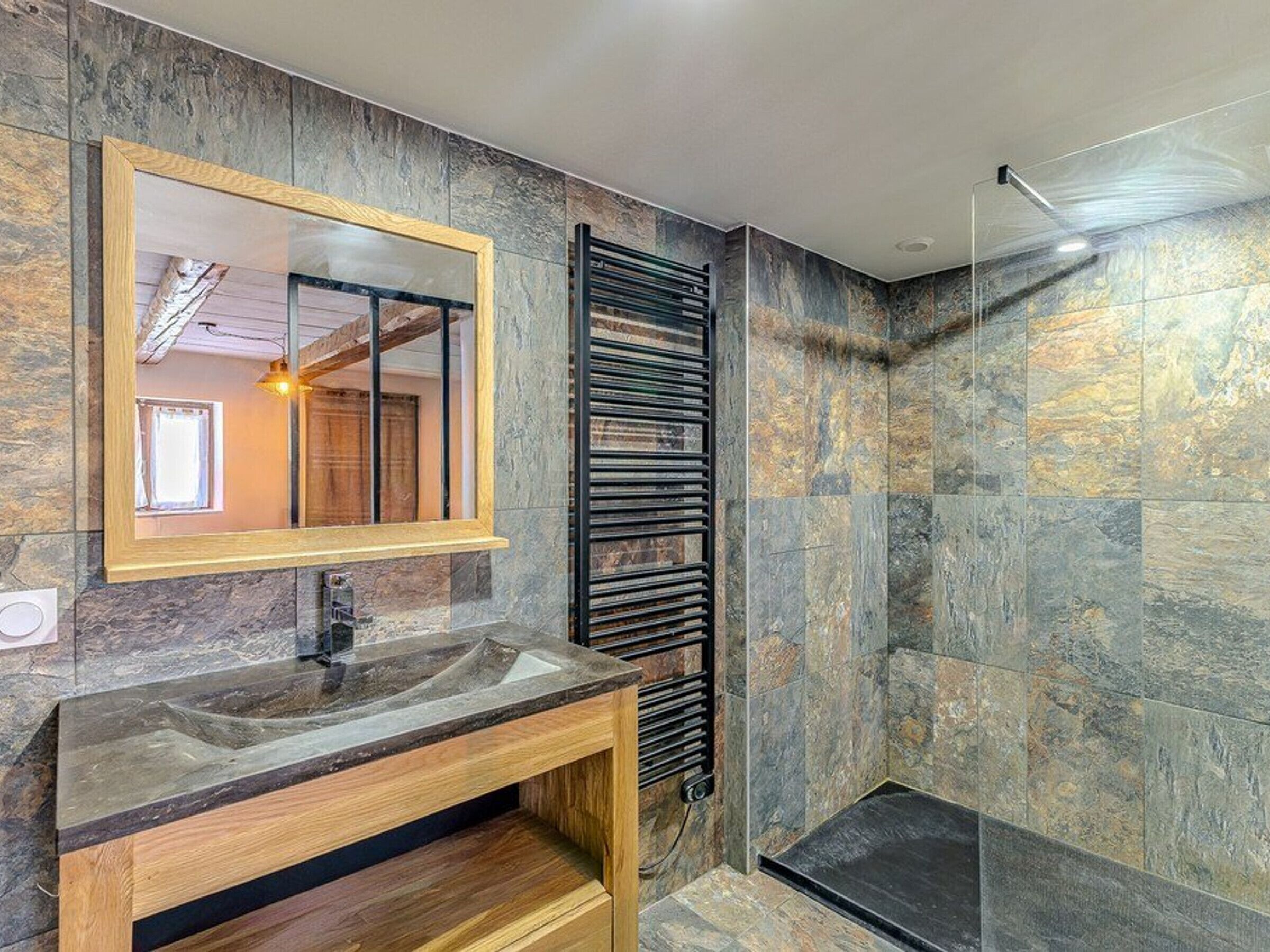 A modern bathroom with a stone wall, a wooden vanity, a black towel rack, and a glass shower enclosure.