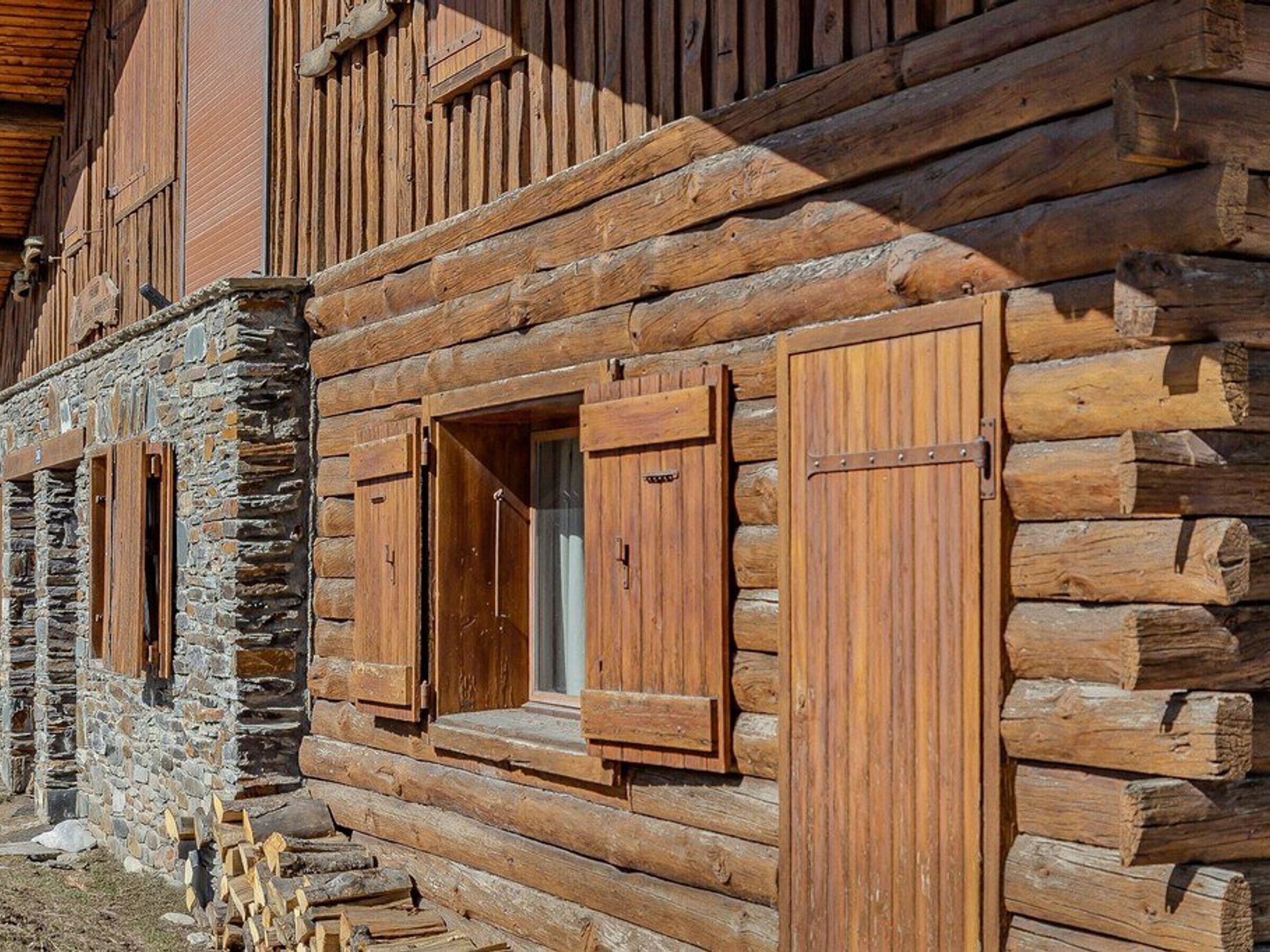 A rustic log cabin with stone foundation, wooden shutters, and a visible window.